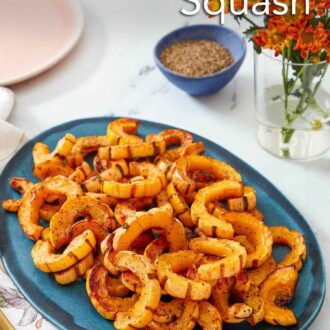 Pinterest graphic of a blue serving platter with pieces of roasted delicata squash with a bowl of pepper and flowers in the background.