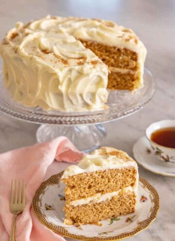 A slice of spice cake on a plate in front of the cut cake on a cake stand.