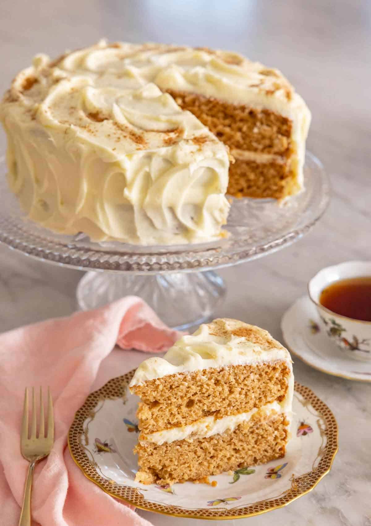 A slice of spice cake on a plate in front of the cut cake on a cake stand.