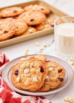 A plate with a couple of white chocolate cranberry cookies with a glass of milk and tray of cookies in the background.