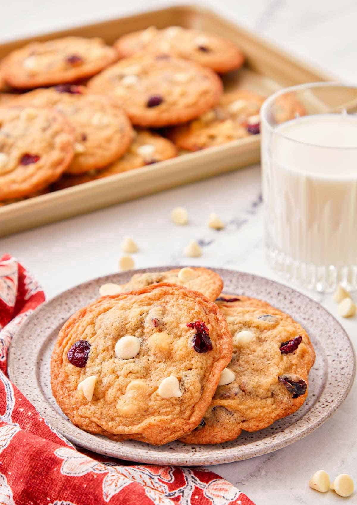 A plate with a couple of white chocolate cranberry cookies with a glass of milk and tray of cookies in the background.