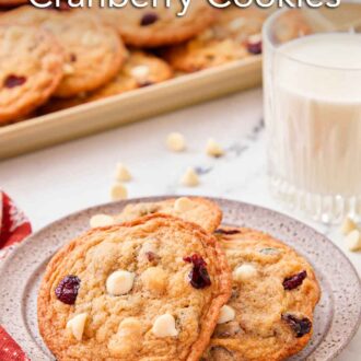Pinterest graphic of a plate with multiple white chocolate cranberry cookies with a glass of milk and a tray of cookies behind it.