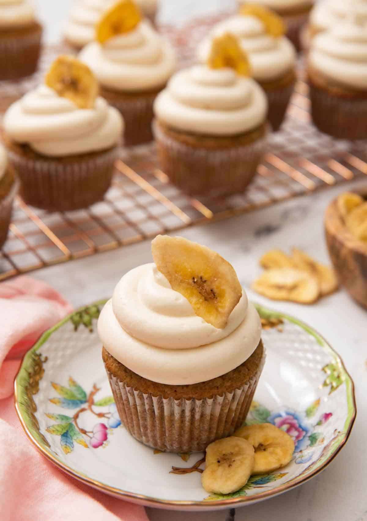 A small plate with a banana cupcake with a banana chip on top and more cupcakes in the back on a cooling rack.