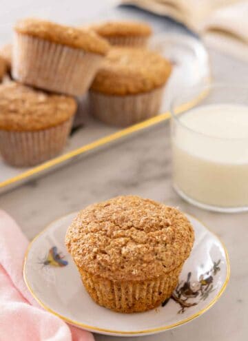 A small plate with a bran muffin with a glass of milk and another platter of muffins in the back.