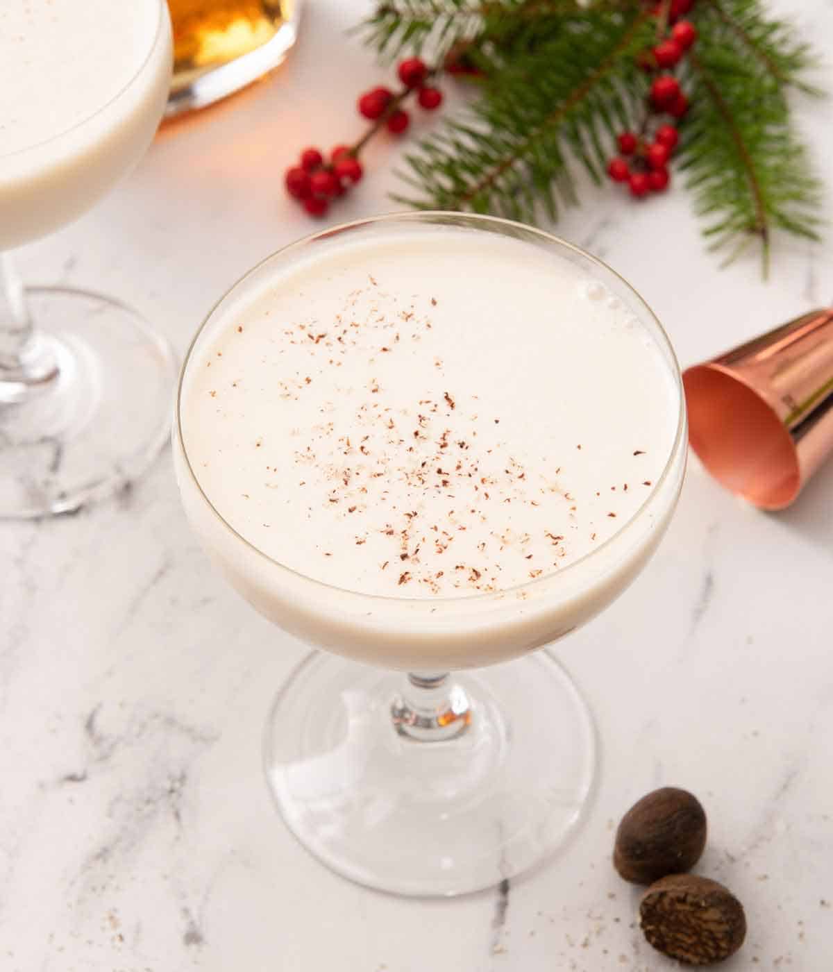 Slightly overhead view of a coupe glass with Brandy Alexander with nutmeg, jigger, and some festive garnish scattered around.