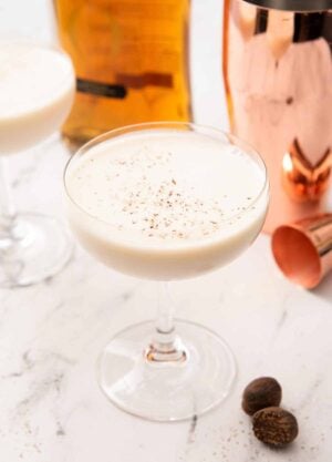 A Brandy Alexander in a coupe glass with fresh nutmeg, a jigger, a shaker, and a bottle of cognac in the back.