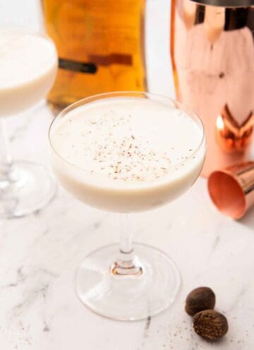 A Brandy Alexander in a coupe glass with fresh nutmeg, a jigger, a shaker, and a bottle of cognac in the back.