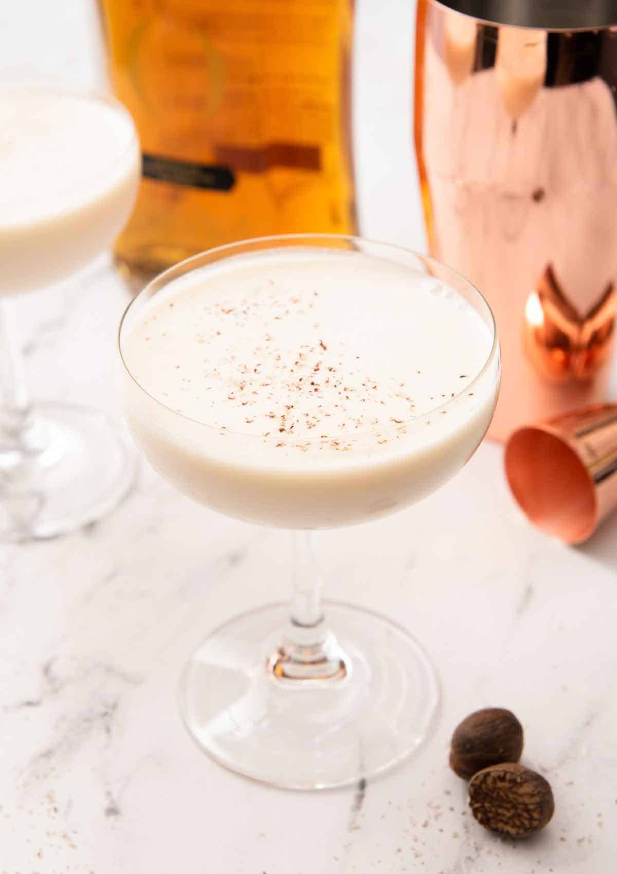 A Brandy Alexander in a coupe glass with fresh nutmeg, a jigger, a shaker, and a bottle of cognac in the back.