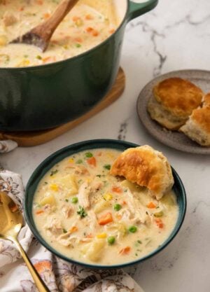 A green bowl of chicken pot pie soup with a biscuit tucked in with more in the background along with the pot.