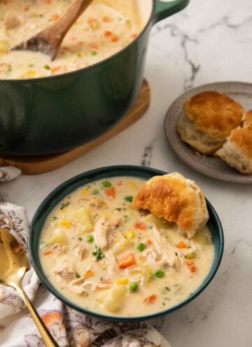 A green bowl of chicken pot pie soup with a biscuit tucked in with more in the background along with the pot.