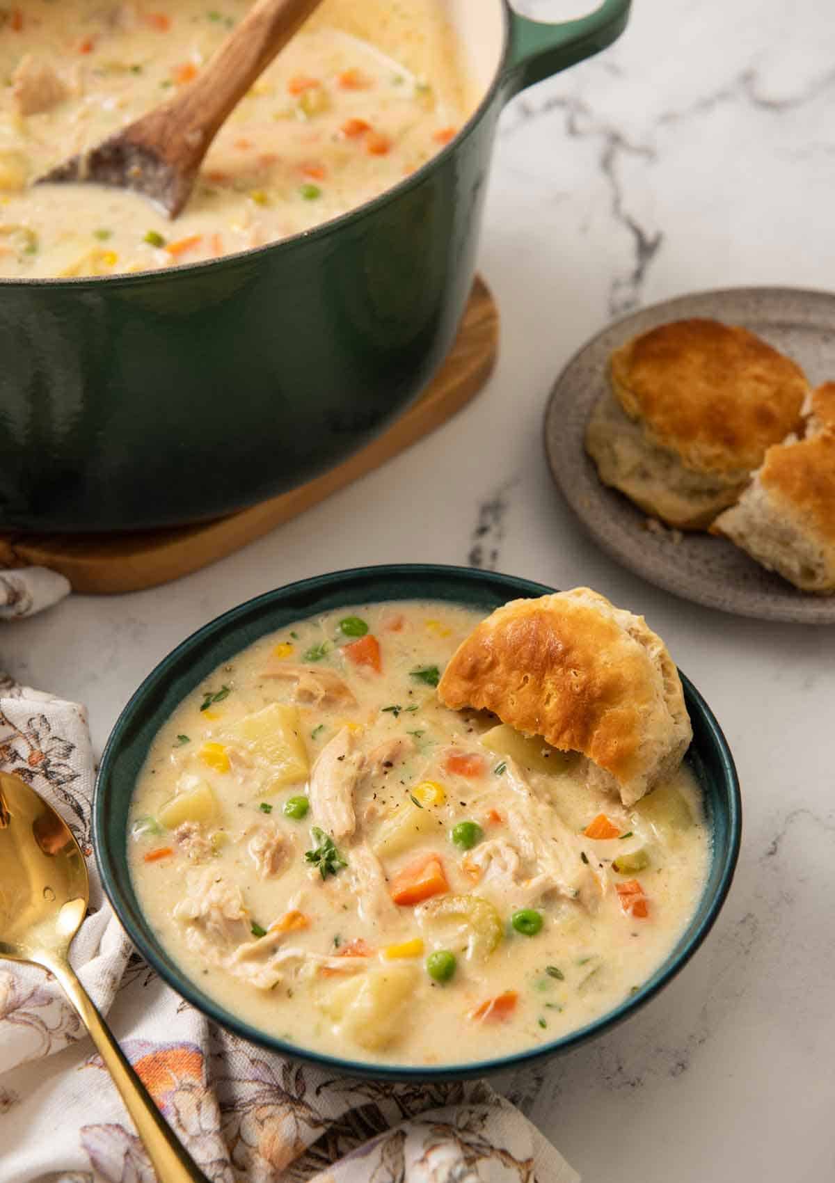 A green bowl of chicken pot pie soup with a biscuit tucked in with more in the background along with the pot.