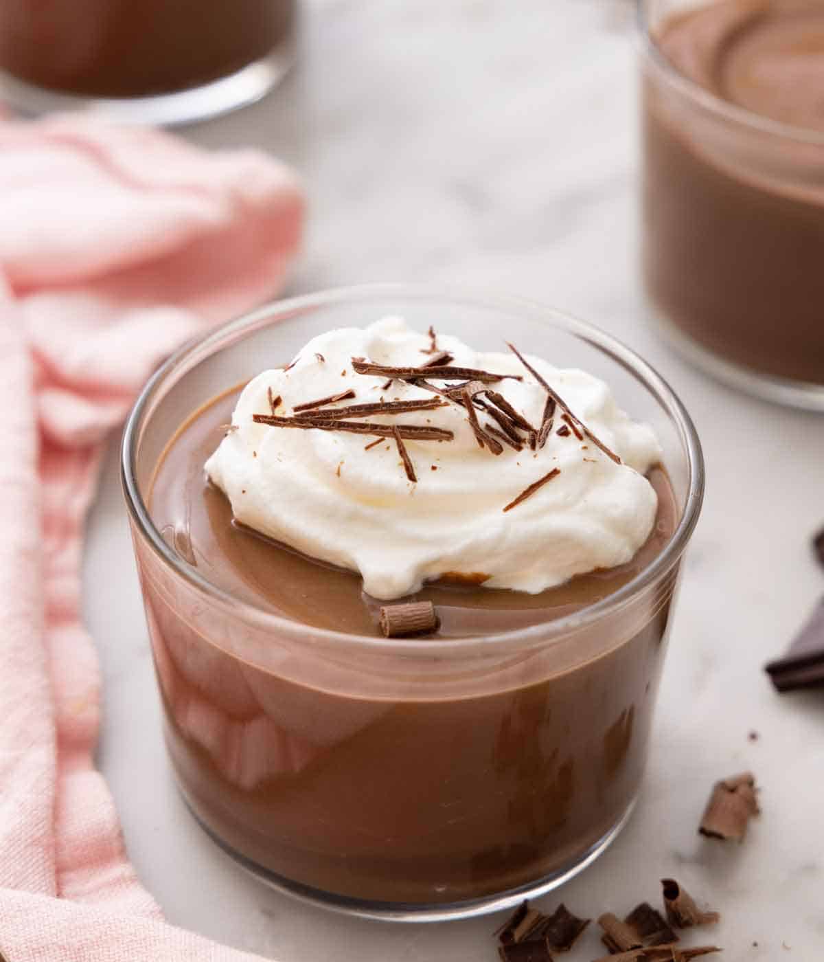 A glass of chocolate pudding with whipped cream on top with shaved chocolate sprinkled on top. Bits of chocolate curls scattered around.