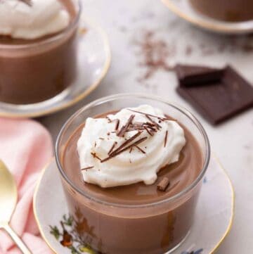 A glass of chocolate pudding with whipped cream and shaved chocolate on top with two more, out of focus, in the background.