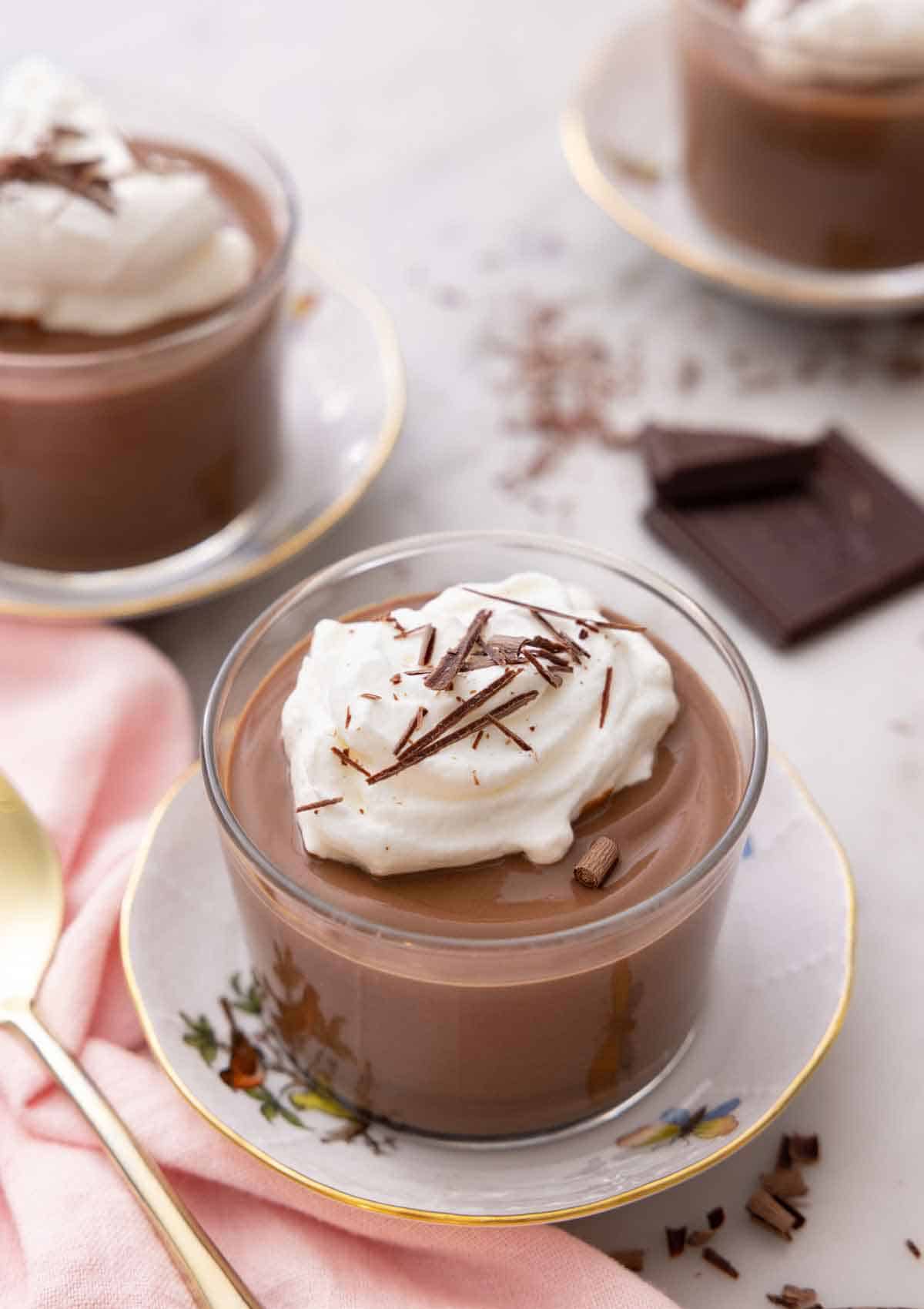 A glass of chocolate pudding with whipped cream and shaved chocolate on top with two more, out of focus, in the background.