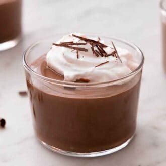 A close up of a glass of chocolate pudding with whipped cream and shaved chocolate on top with two more glasses and pieces of chocolate behind it.