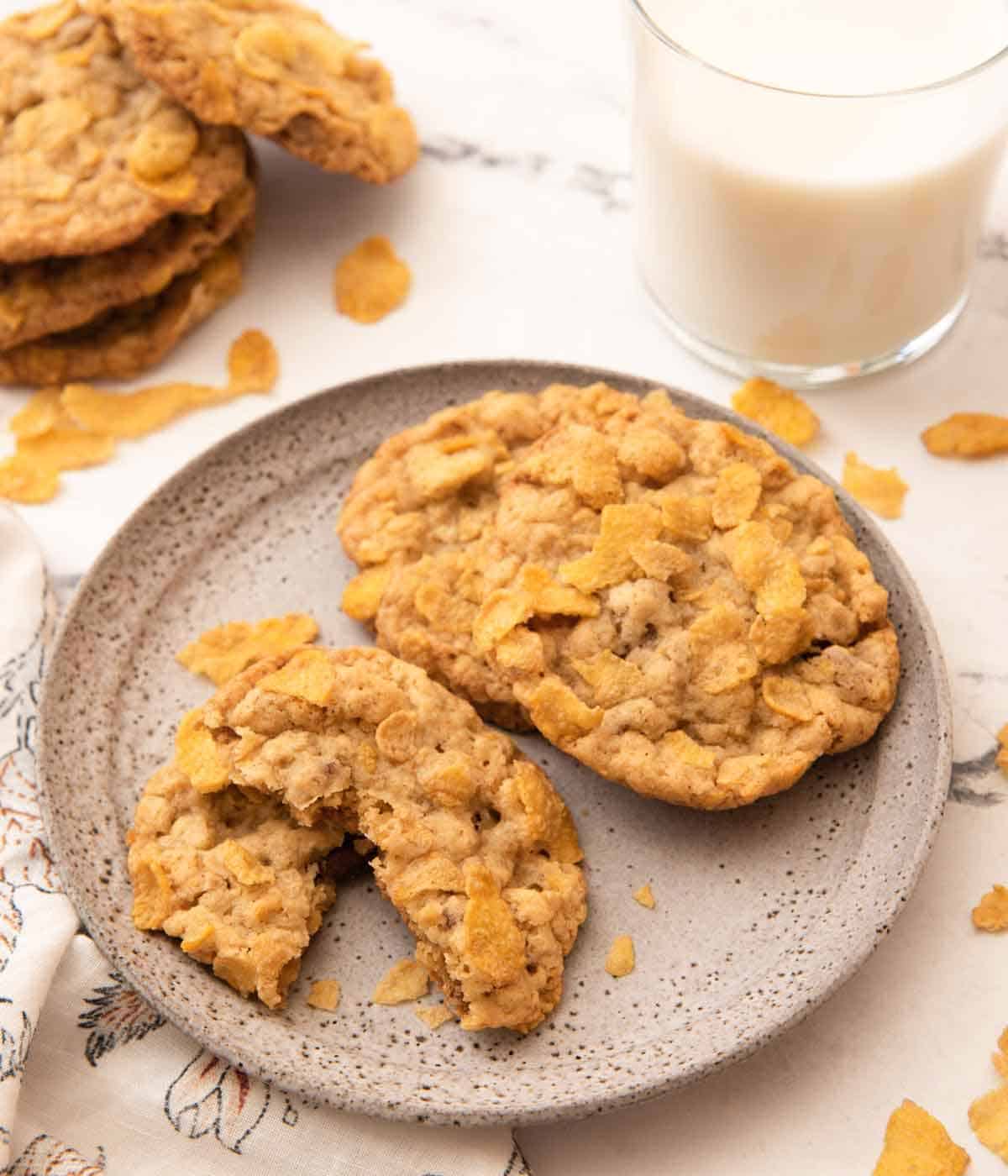 A plate with multiple cornflake cookies with one torn in half with a stack in the background.