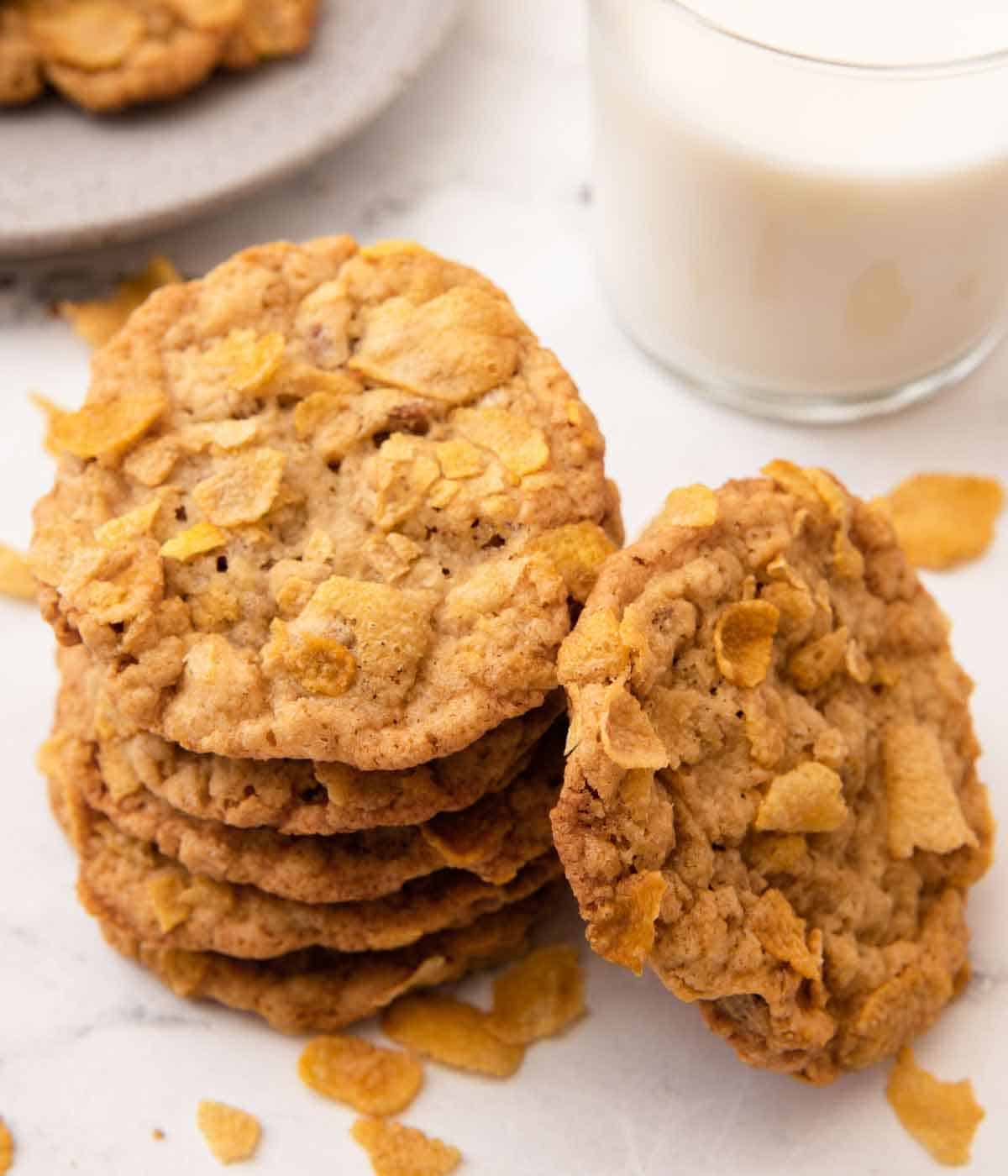 A stack of five cornflake cookies with one leaning beside it with cornflakes scattered around.