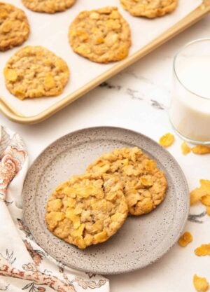A plate with two cornflake cookies with a sheet pan with more and a glass of milk behind the plate.