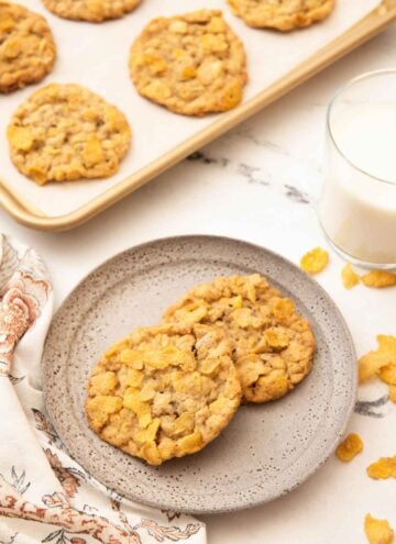 A plate with two cornflake cookies with a sheet pan with more and a glass of milk behind the plate.