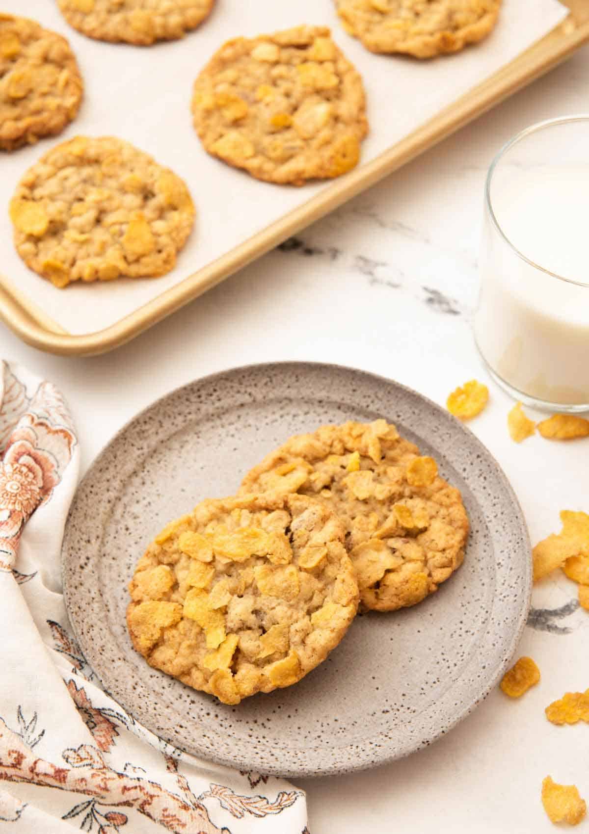 A plate with two cornflake cookies with a sheet pan with more and a glass of milk behind the plate.