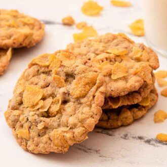 A small stack of three cornflake cookies with one leaning against it with more cookies in the back and cornflakes scattered around.