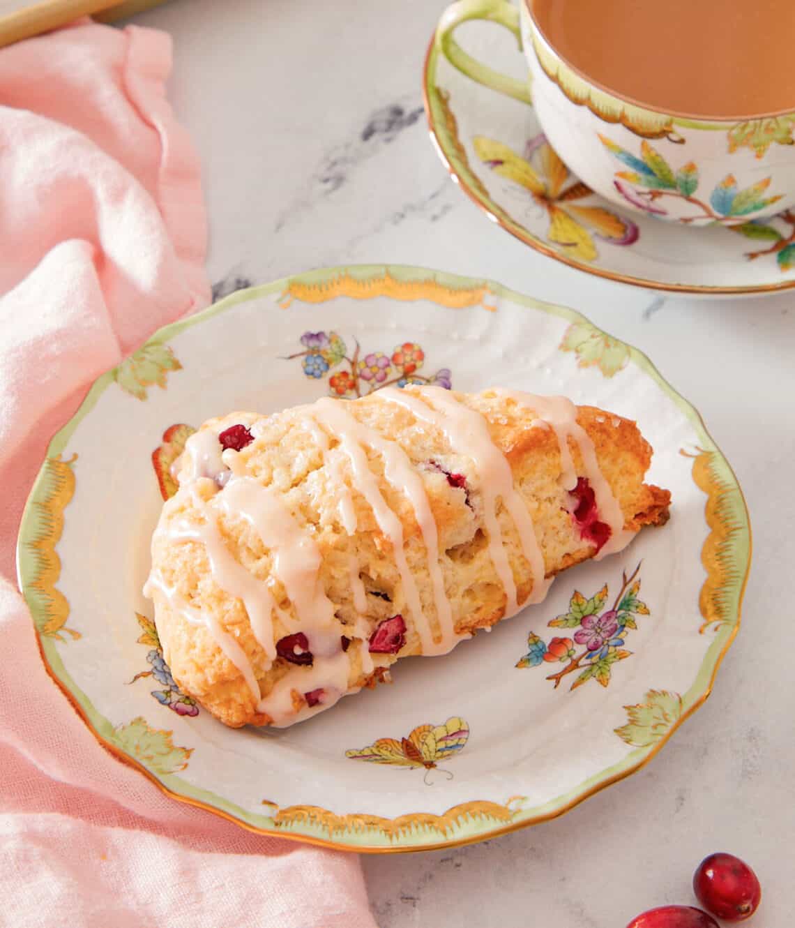A plate with a cranberry orange scone with a drizzle of glaze over top with a mug of coffee in the back.