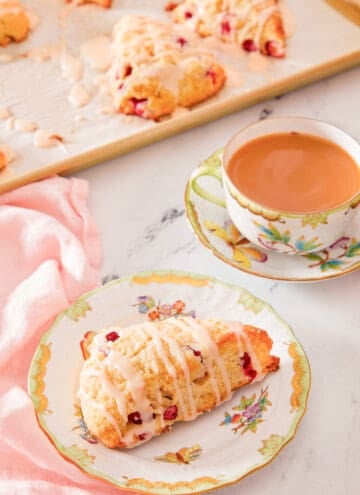 A plate with a cranberry orange scone with a mug of coffee in the back with more scones on a baking sheet behind it.
