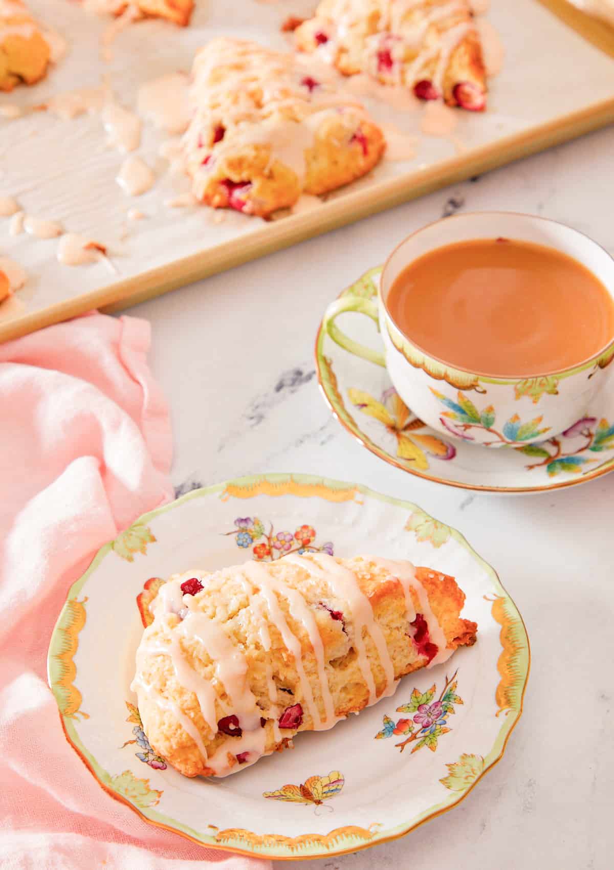 A plate with a cranberry orange scone with a mug of coffee in the back with more scones on a baking sheet behind it.