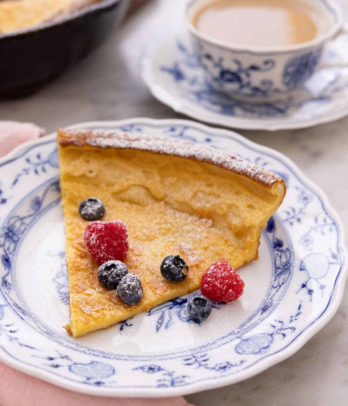 A plate with a slice of Dutch baby with powder sugar and fresh berries on top with a cup of tea in the back.
