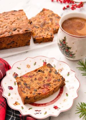 A festive plate with a slice of fruit cake with a mug of coffee and more cake behind it.
