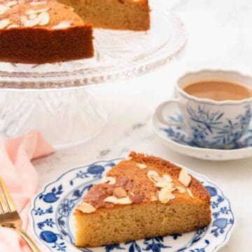 A plate with a slice of honey cake with the rest of the cake and a coffee in the background.