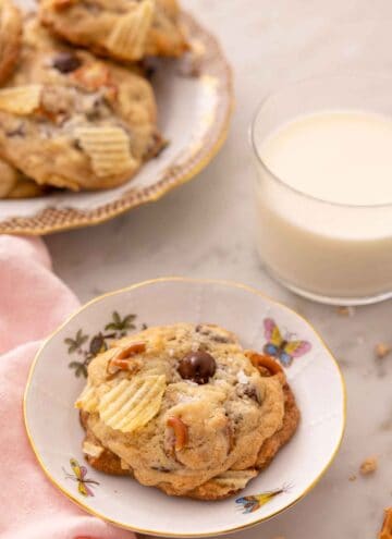 A small plate with a kitchen sink cookie with a glass on milk and a platter of more cookies behind it.