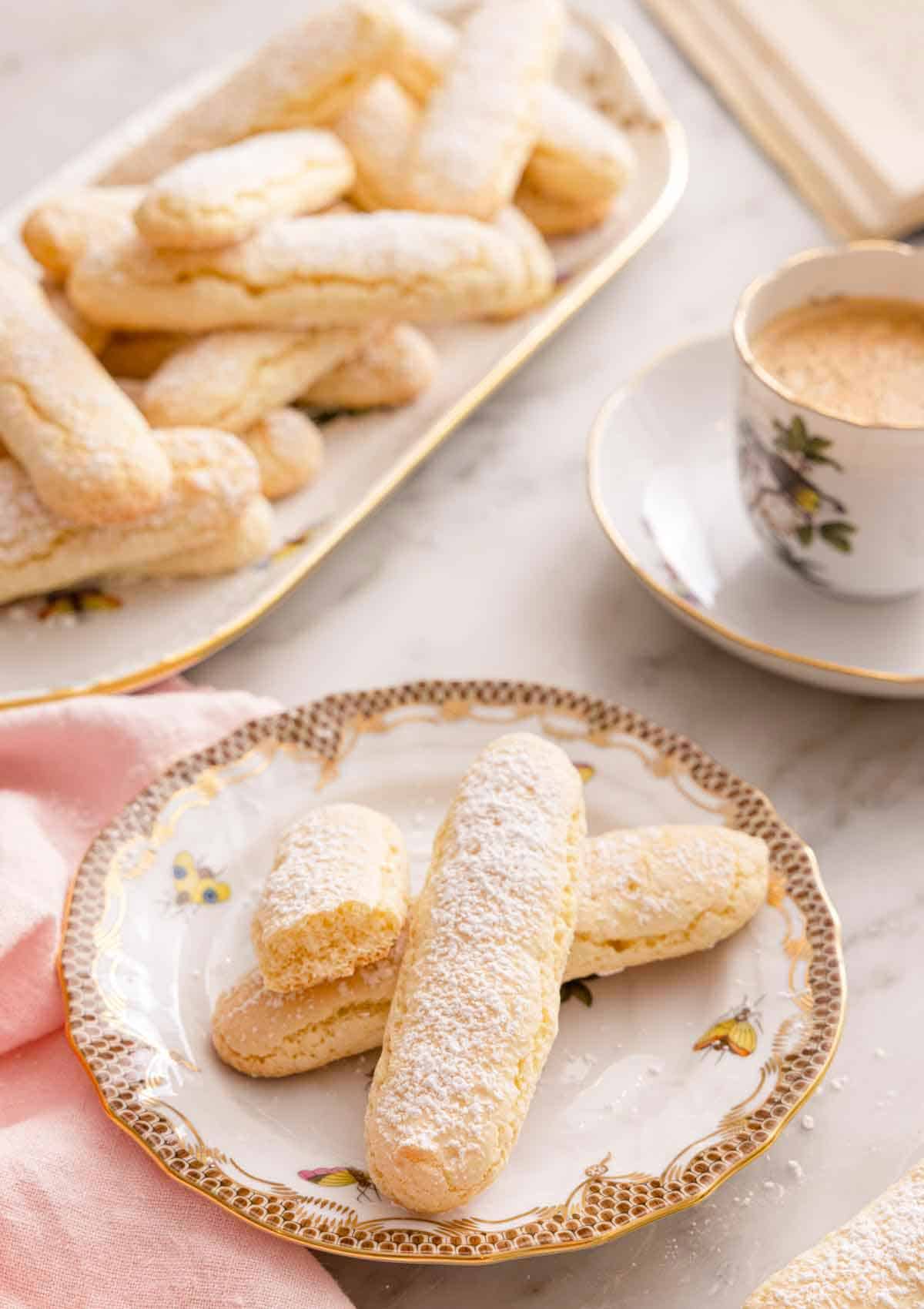 A plate with three ladyfingers with a cup of coffee and a tray of additional ladyfingers in the back.