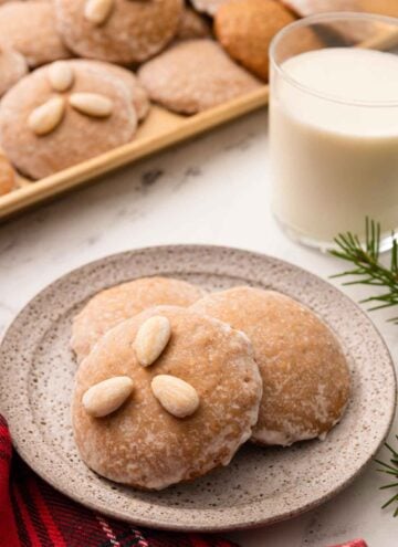 A plate with three Lebkuchen with one with three almonds on top with a glass of milk in the back and more cookies.