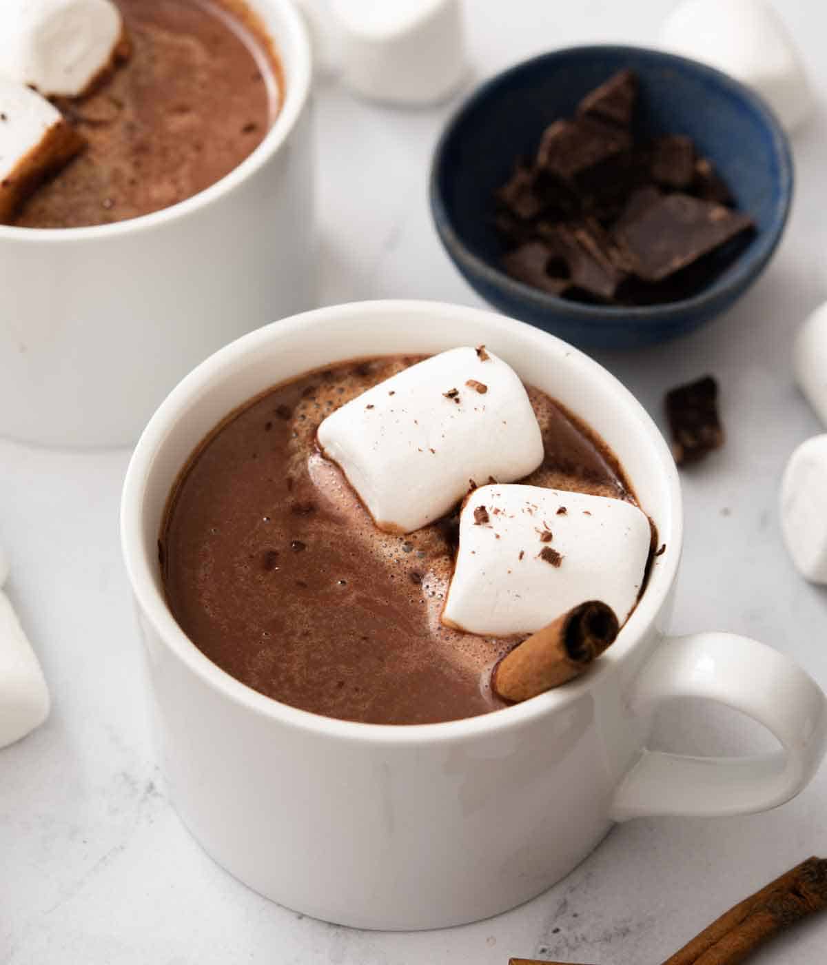 A white mug with Mexican hot chocolate topped with two marshmallows and a cinnamon stick with a bowl of chocolate in the back.