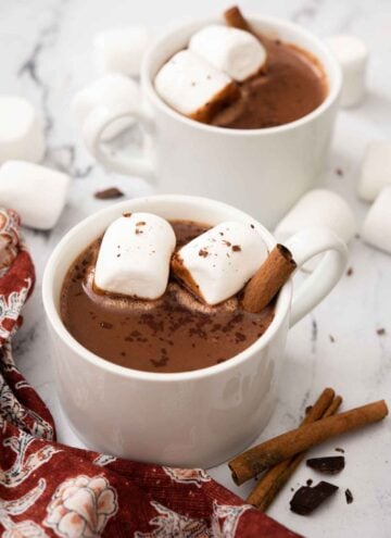 Two mugs of Mexican hot chocolate with marshmallows and cinnamon sticks inside. More garnish scattered around the counter.