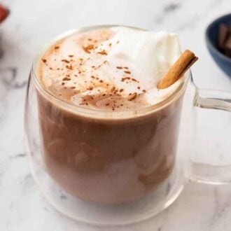 A clear glass of Mexican hot chocolate with whipped cream and cinnamon stick on top with a small bowl of chocolate in the back.