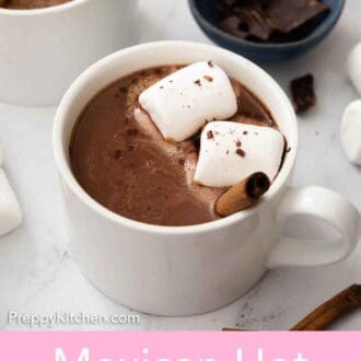 Pinterest graphic of a mug of Mexican hot chocolate with marshmallows and a cinnamon stick on top with a small bowl of chocolate in the back.