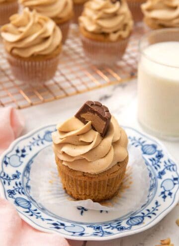 A small plate with a peanut butter cupcake with the liner pulled down and glass of milk and more cupcakes in the back.