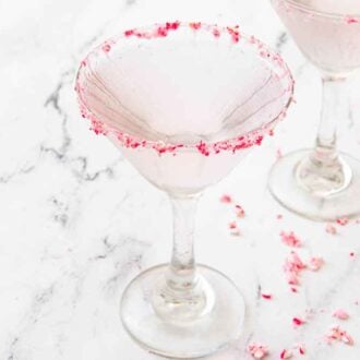 A glass of peppermint martini with crushed peppermint candies on the rim with a second one behind it and crushed candies scattered around.