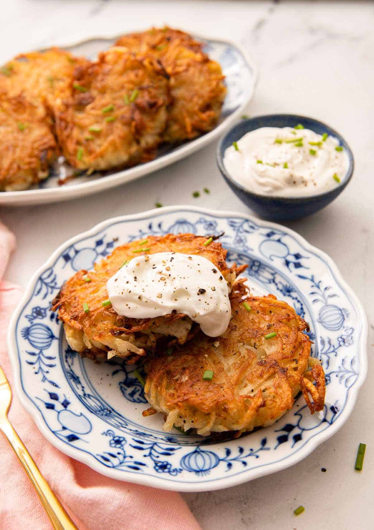 A plate with two potato pancakes with a dollop of sour cream on top. More pancakes and dip in the background.