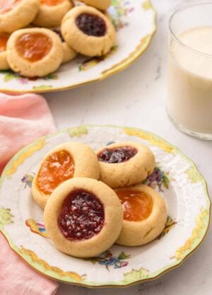 A plate with four thumbprint cookies with a glass of milk and another plate of cookies in the back.
