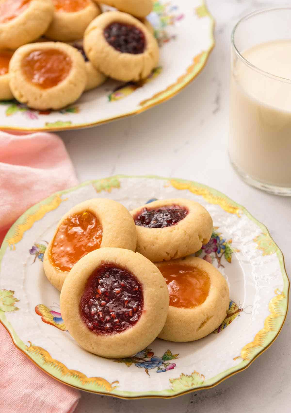 A plate with four thumbprint cookies with a glass of milk and nothing plate of cookies in the back.