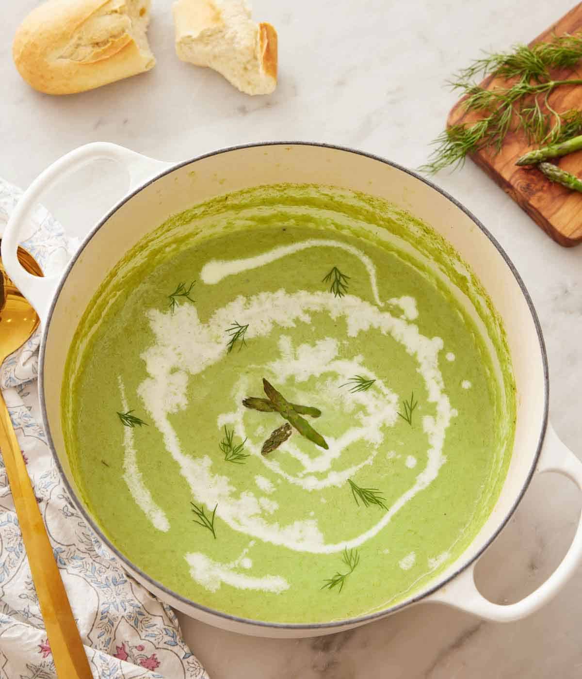Overhead view of a pot of asparagus soup with some fresh dill and asparagus as garnish and a drizzle of cream.
