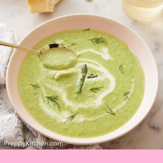 Pinterest graphic of a bowl of asparagus soup with a spoonful being lifted out of the bowl. Some bread and wine in the background.