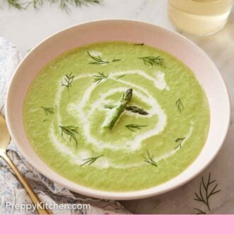 Pinterest graphic of a bowl of asparagus soup with fresh dill and asparagus as garnish and a glass of wine in the back.