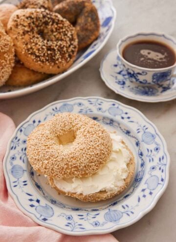 A plate with a sliced bagel with sour cream smeared inside. A cup of coffee and more bagels in the back.