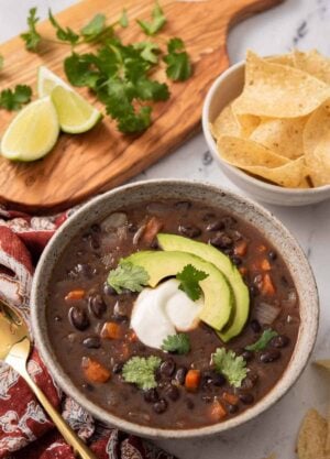 A bowl of black bean soup with sour cream and sliced avocado as garnish. A bowl of chips, lime wedges, and cilantro in the back.