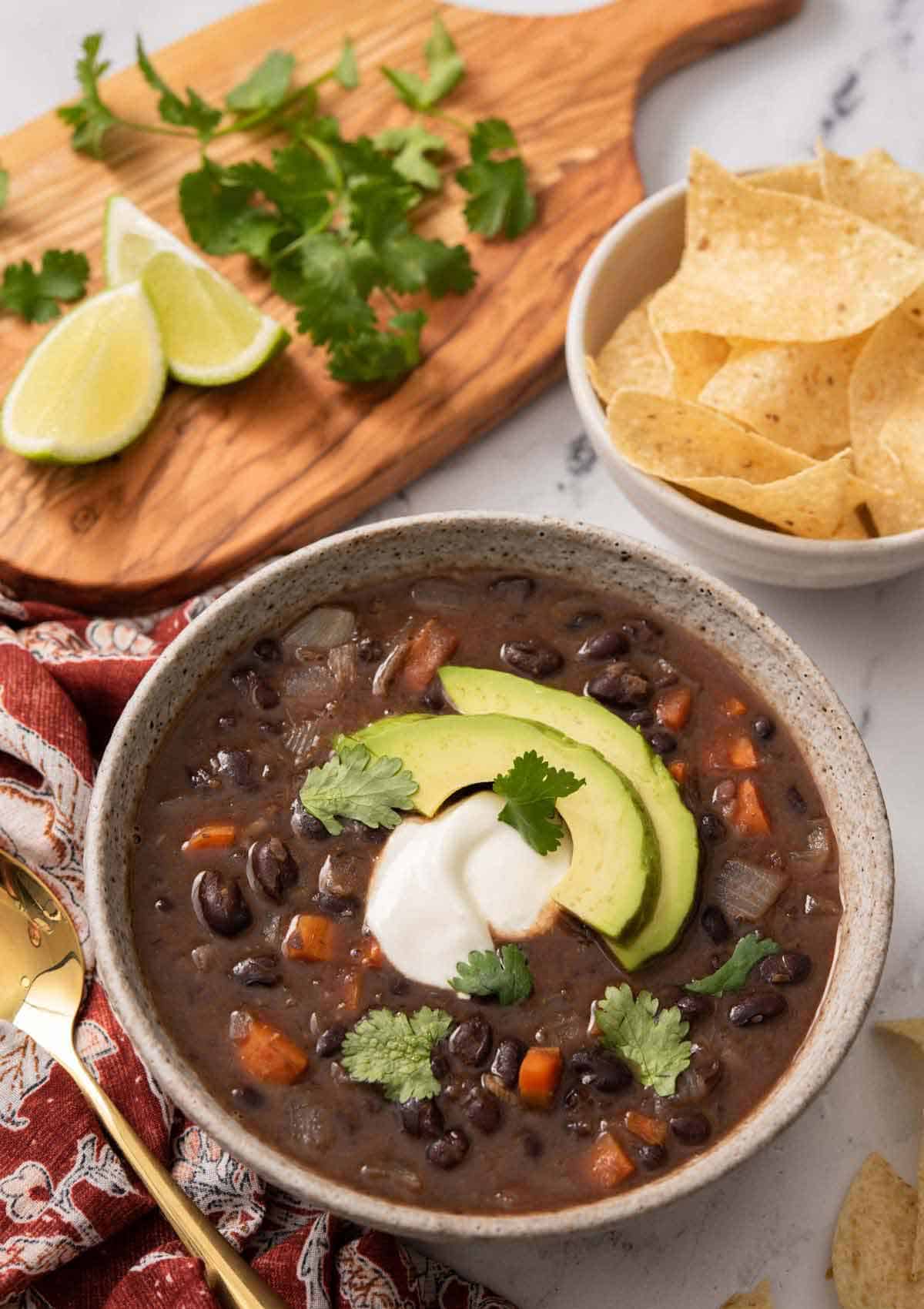 A bowl of black bean soup with sour cream and sliced avocado as garnish. A bowl of chips, lime wedges, and cilantro in the back.