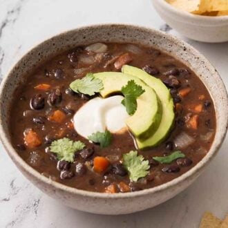 A bowl of black bean soup with sliced avocado, sour cream, and cilantro on top. Lime wedges and tortilla chips scattered around.